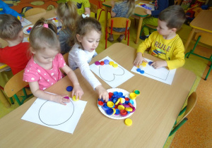 Dzieci układają nakrętki plastikowe na szablonie figur geometrycznych.